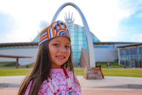 A child wearing traditional Native American dress smiles in front of the arched architectural entry of the First American Museums in Oklahoma.