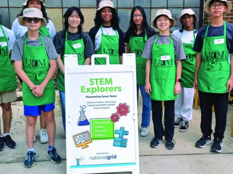 A group of about eight young volunteers stand next to a sign that says STEM Explorers. All of them are wearing bright green aprons.