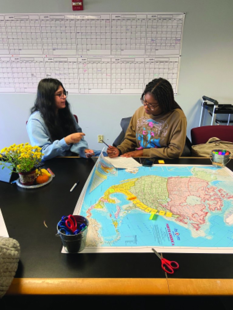 Two young people sit at a table with a map stretched in front of them.