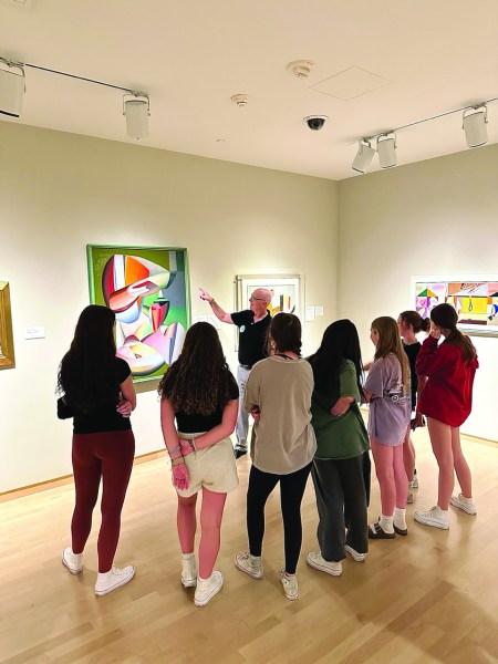 A group of students stand listening to a docent explain a painting hanging on the wall. 