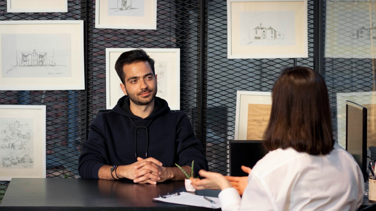 A man sits on one side of a table facing a woman who is interviewing him. The room has a number of framed prints on the wall behind the man.