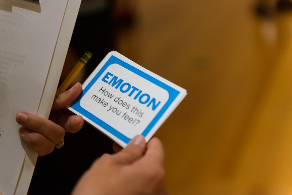 A closeup image of a teacher’s hand holding a pencil and a card that reads “Emotion: How does this make you feel?”