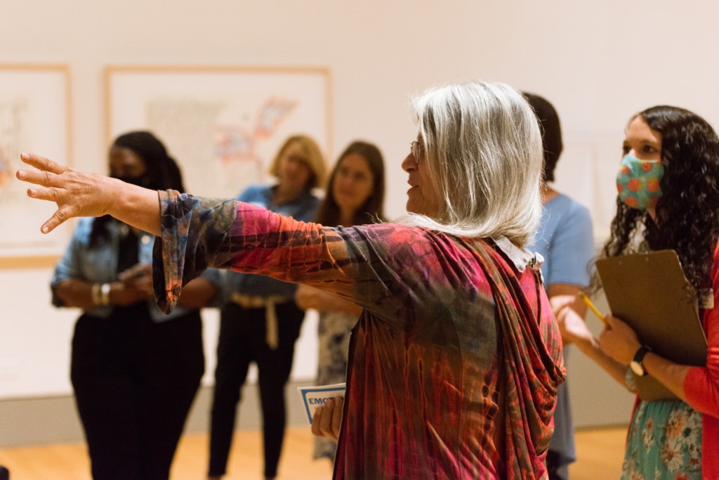 A group of teachers are gathered in the gallery during a workshop as one points to an image and describes how it relates to the emotion card in her hand.