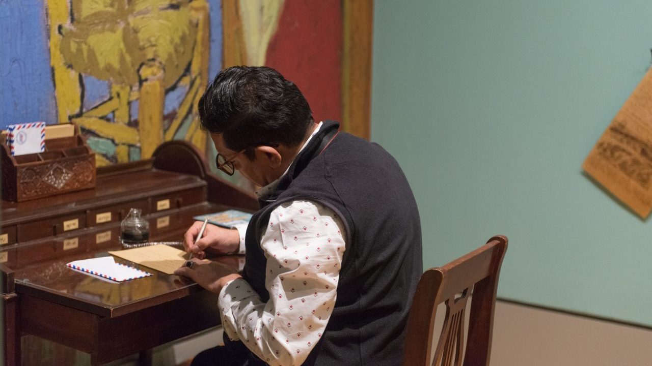A visitor seated at an old-fashioned writing desk writes a letter within an exhibition on the art and influence of Vincent Van Gogh.