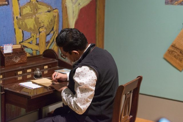 A visitor seated at an old-fashioned writing desk writes a letter within an exhibition on the art and influence of Vincent Van Gogh.