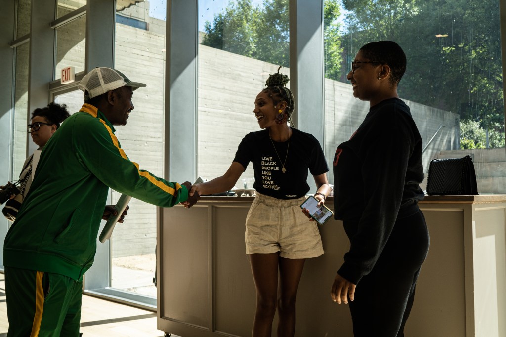 Two museum staff members greeting and shaking hands with a visitor in front of a large window.