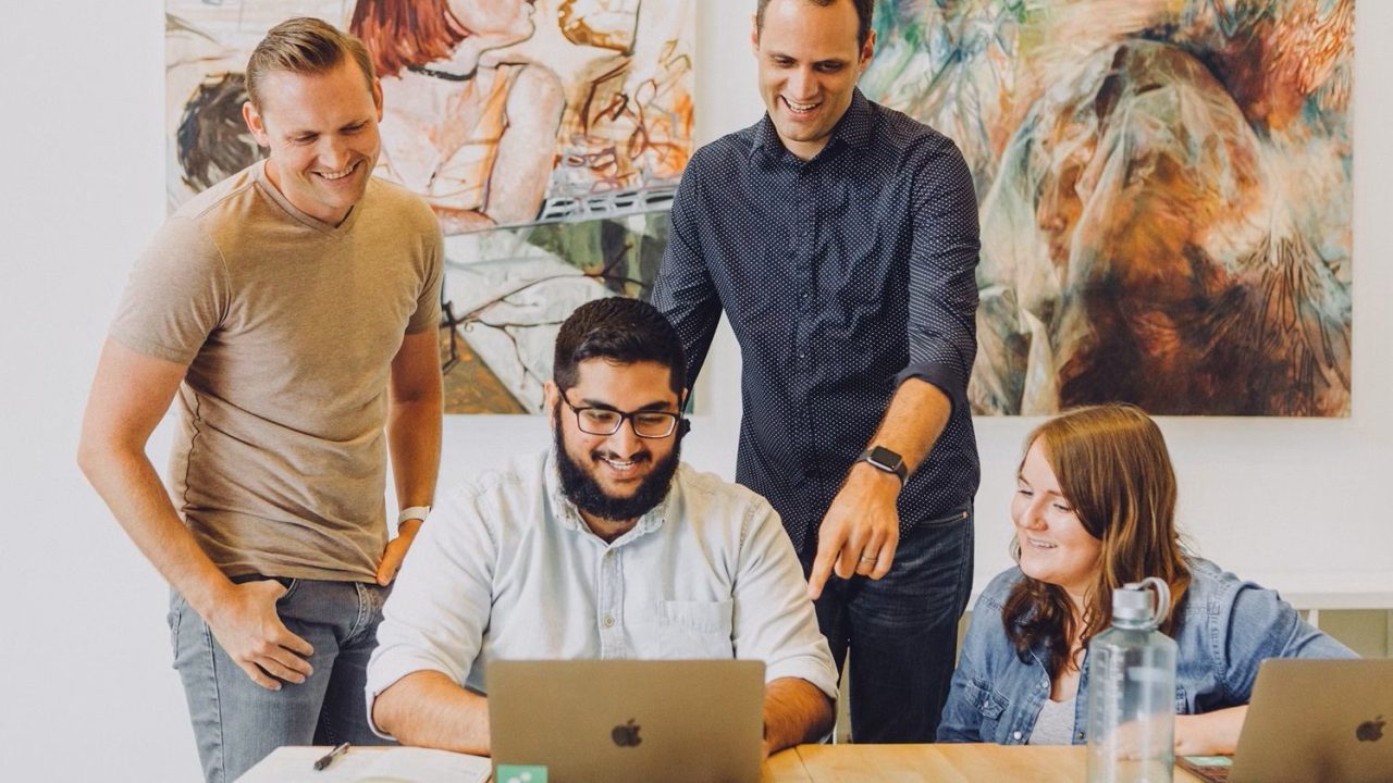 A group of four people stand and sit in front of a laptop with a colorful painting in the background.