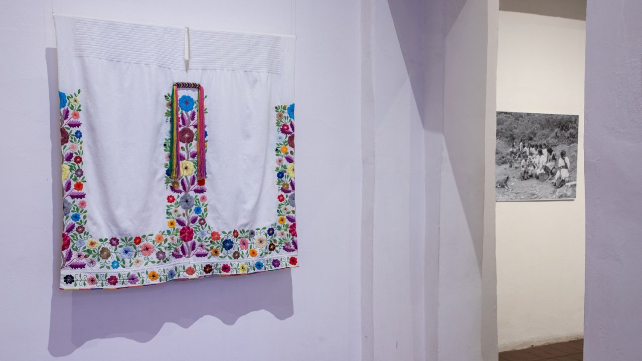 An embroidered shall hangs on a white wall in the gallery.