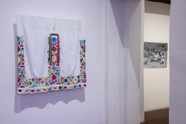 An embroidered shall hangs on a white wall in the gallery.