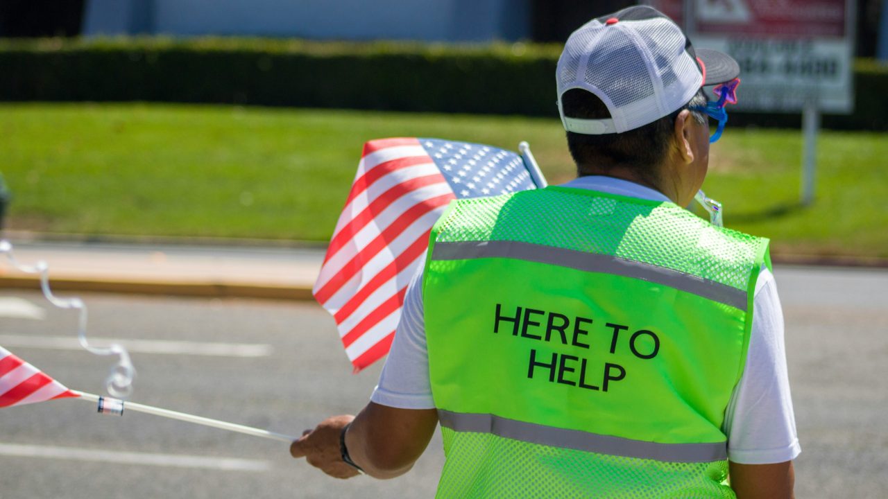 A person standing by a street wearing a neon vest reading "Here to Help."
