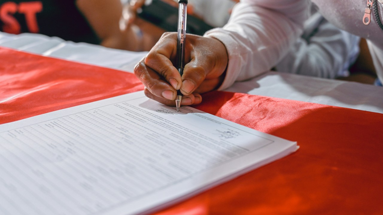 A person's arm signs a document in front of them that is sitting on a red flag.