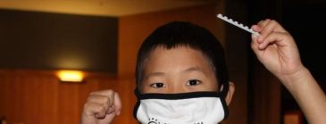 A young boy sits at a table with a Lego creation in front of him holding up a Lego piece in his hand. He is wearing a facemask over his mouth. 