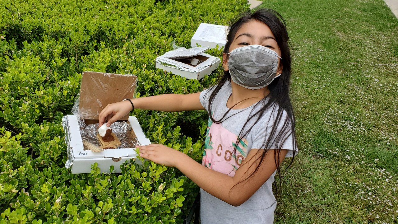 A young girl stands next to a hedge wearing a facemask covering her mouth.
