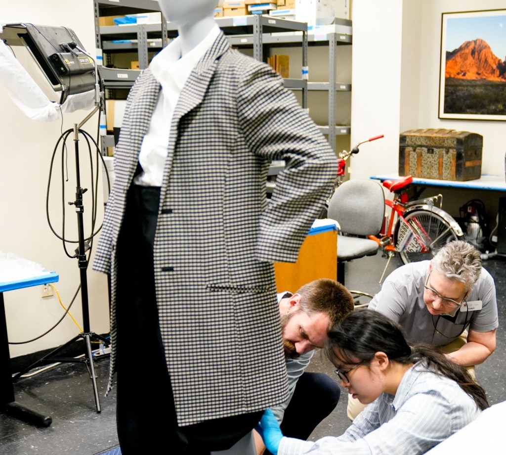 A group of people inspecting the leg of a pair of pants on a dressed mannequin