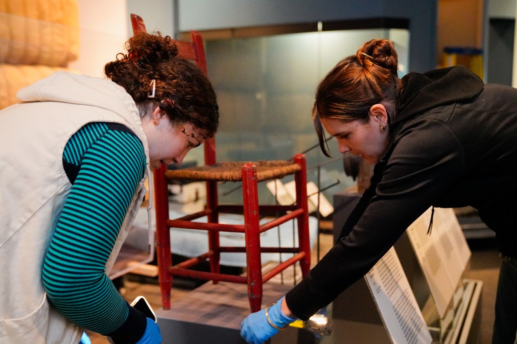 Two people wearing gloves and working on an antique chair on a pedestal