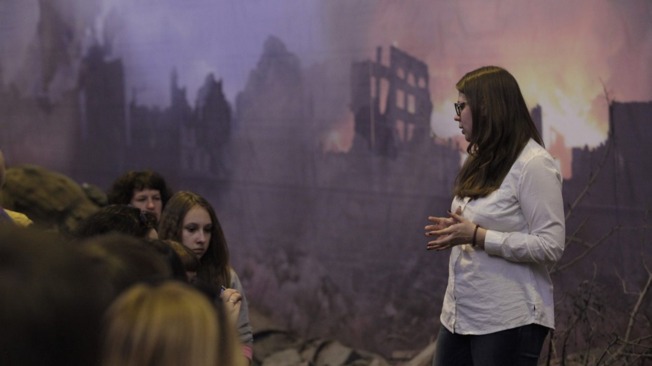 An adult addressing a seated group of young people in front of a large backdrop depicting a burning building