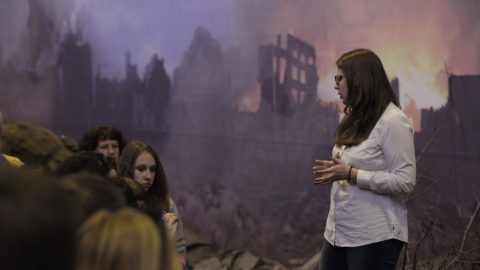 An adult addressing a seated group of young people in front of a large backdrop depicting a burning building