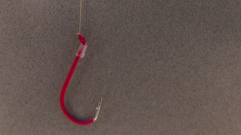 A red fish hook dangles from a clear fishing line in front of a dark gray background.