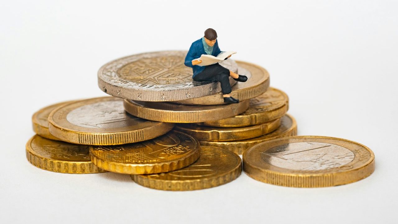 A miniature figure reading a book sits atop a large stack of gold coins