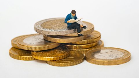 A miniature figure reading a book sits atop a large stack of gold coins