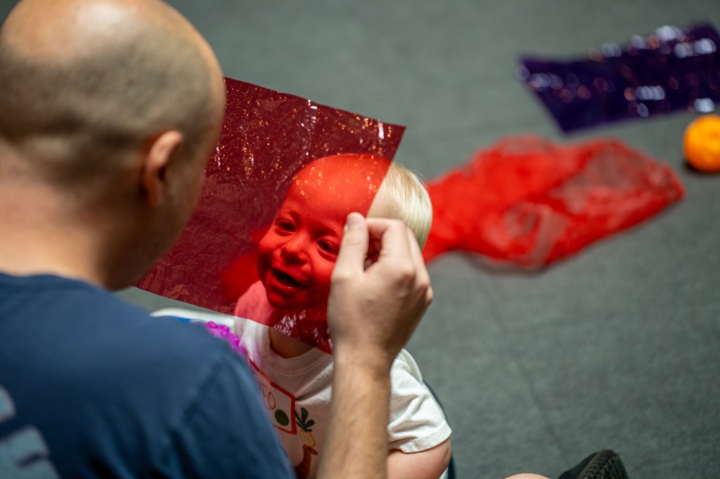 A small child gazes up at an adult holding a transparent red square of plastic