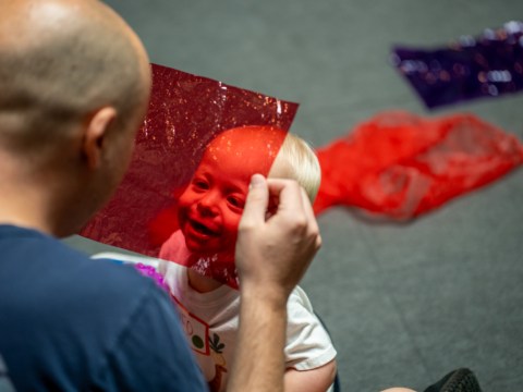A small child gazes up at an adult holding a transparent red square of plastic