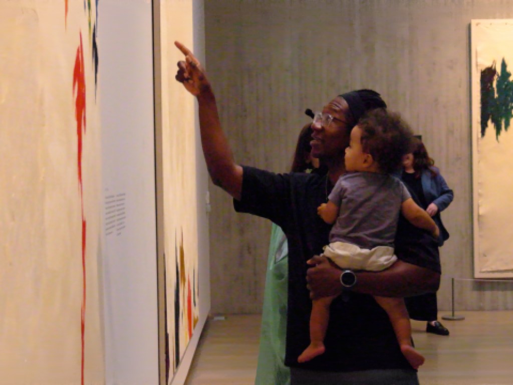 An adult holds a child while pointing at a painting, both looking at the artwork