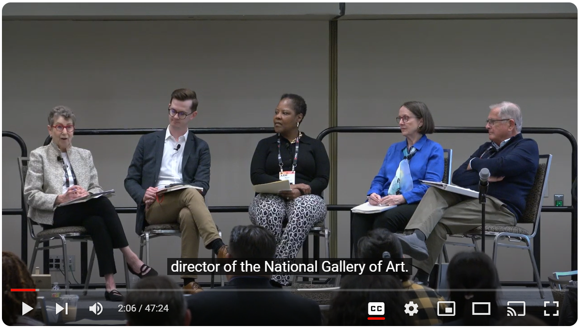 Screenshot of the Imprint Series: Museums For a New Century: 1984, Today, and Tomorrow video with four panelists seated on a dais in front of a crowd of tables and chairs.