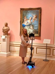 A guide speaking into a device mounted on a wheeled tripod in front of a painting in a gallery