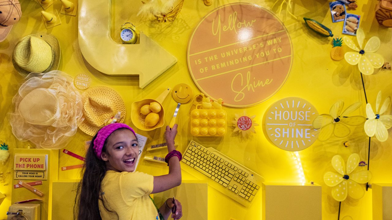 A young person stands in front of a vibrant yellow wall adorned with various yellow objects attached to it.