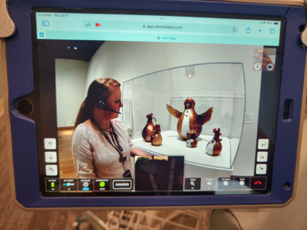 A tablet screen showing a camera feed of a guide in front of a display case of sculptures