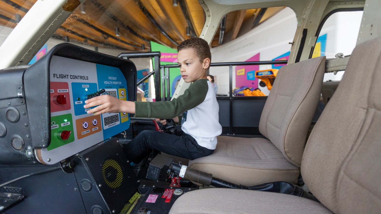 A child sitting in a mock cockpit adjusting a knob that reads "flight controls" next to an indicator screen labeled "patient health monitor."