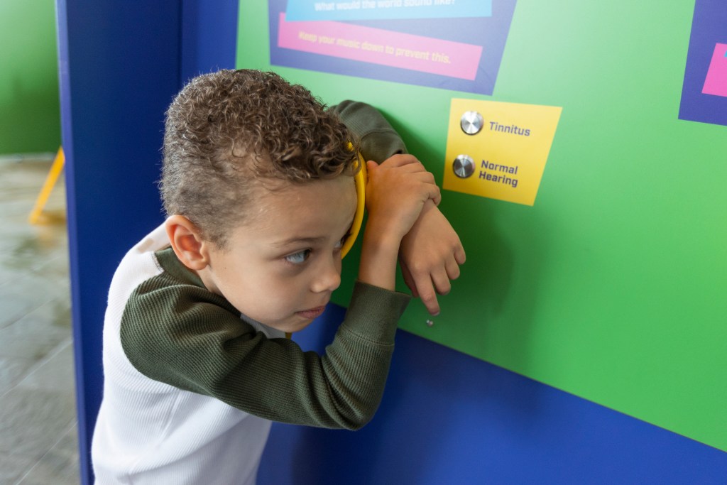 A child leaning in to listen to a speaker next to a control panel with buttons labeled "tinnitus" and "normal hearing."
