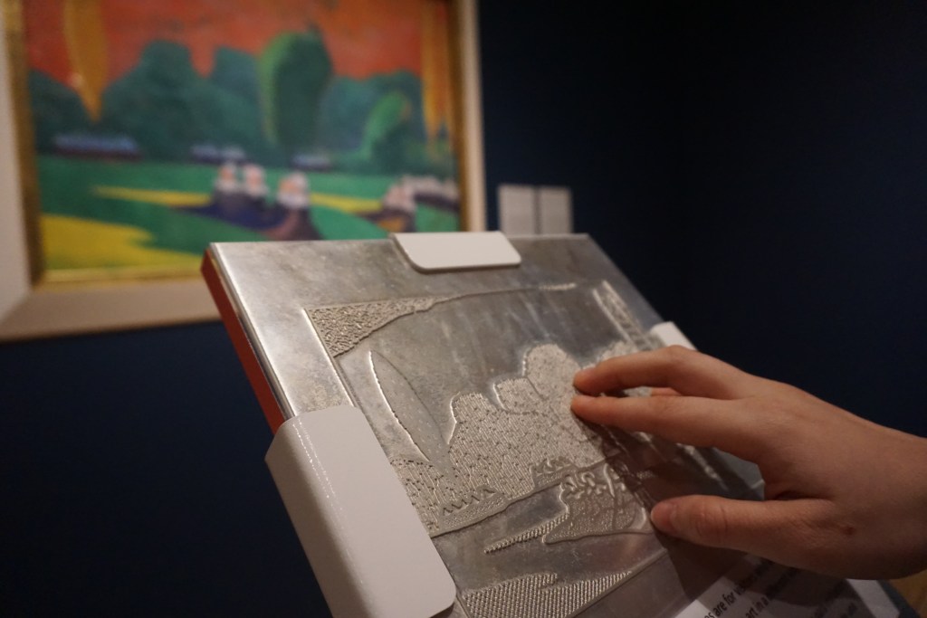 A metal plaque in front of a painting showing an rendering of the painting in raised lines, with a person touching the lines.