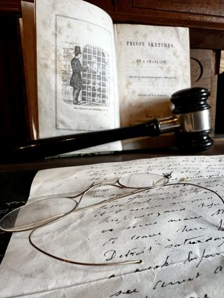 A closeup of a handwritten note with a pair of wire rimmed glasses sit on a table where an open book sits with a judge's gavel in front.