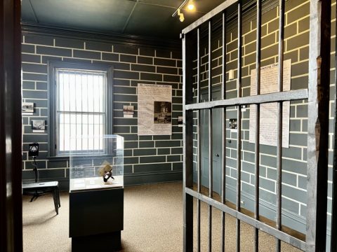 The inside of a jail cell with the iron bar door open and several text panels displayed on the wall with a cased object in the center of the room.