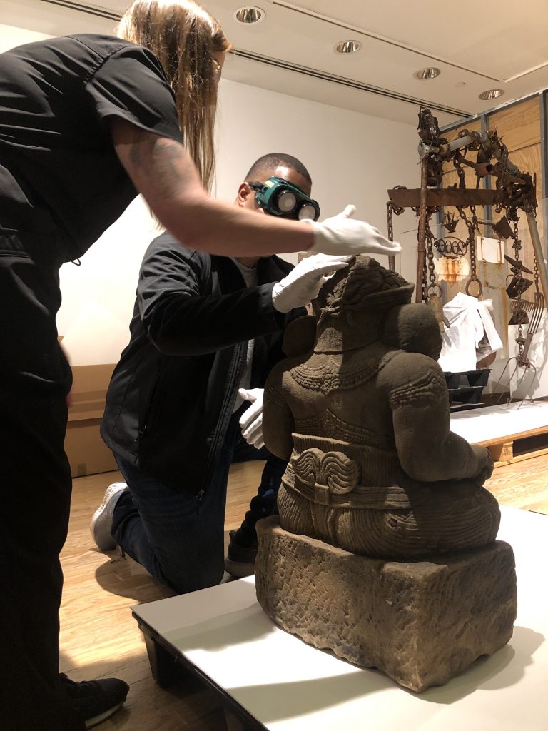 A person wearing goggles and touching a sculpture with gloves, while another person without goggles guides them
