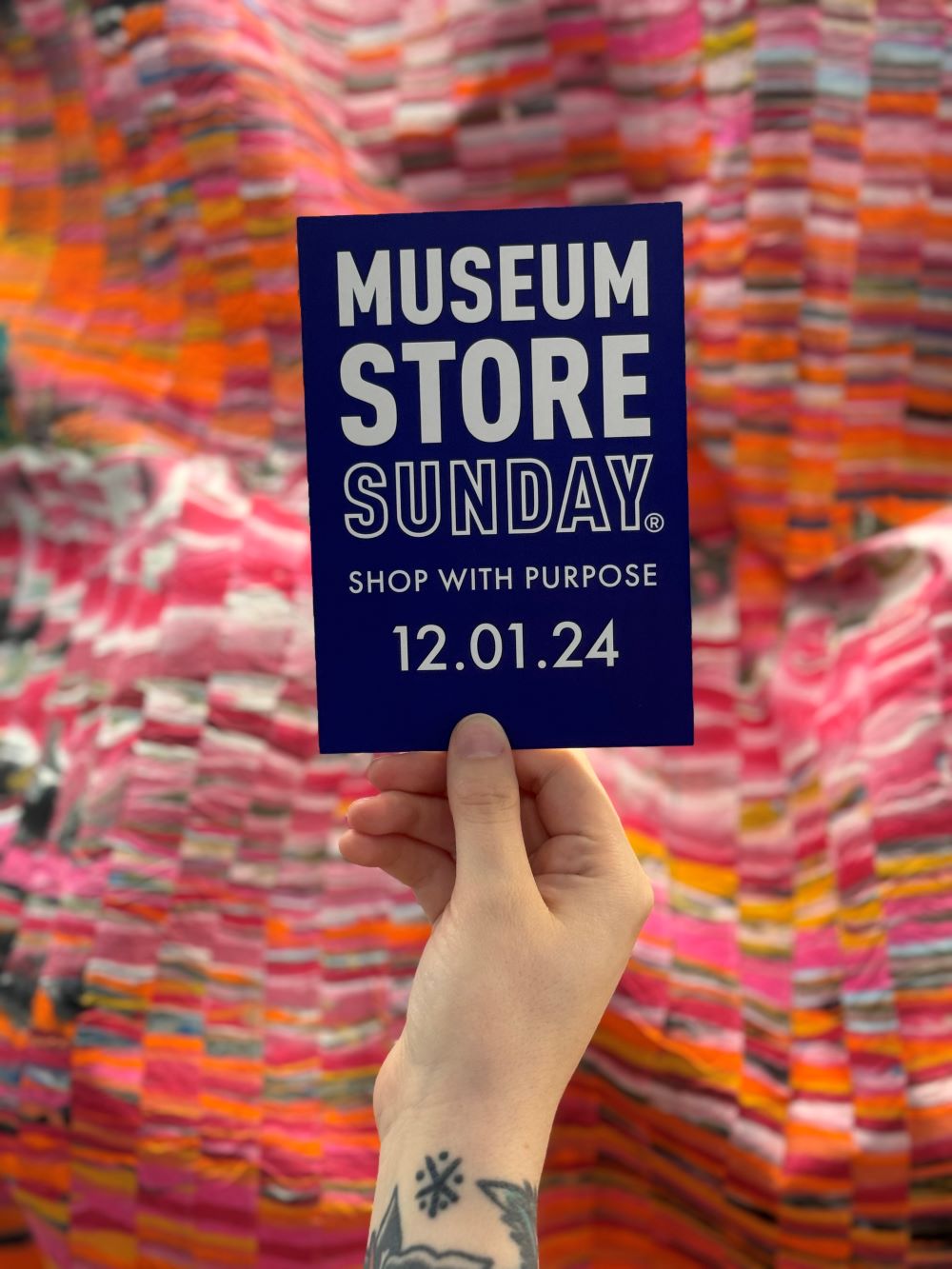 A person holding up a flyer that reads "Museum Store Sunday / Shop With Purpose / 12.01.24"