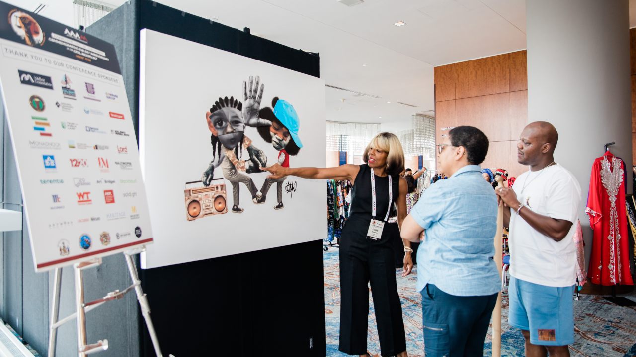 Two people in a convention center listening to a person speaking and pointing at a poster of a collage-style artwork