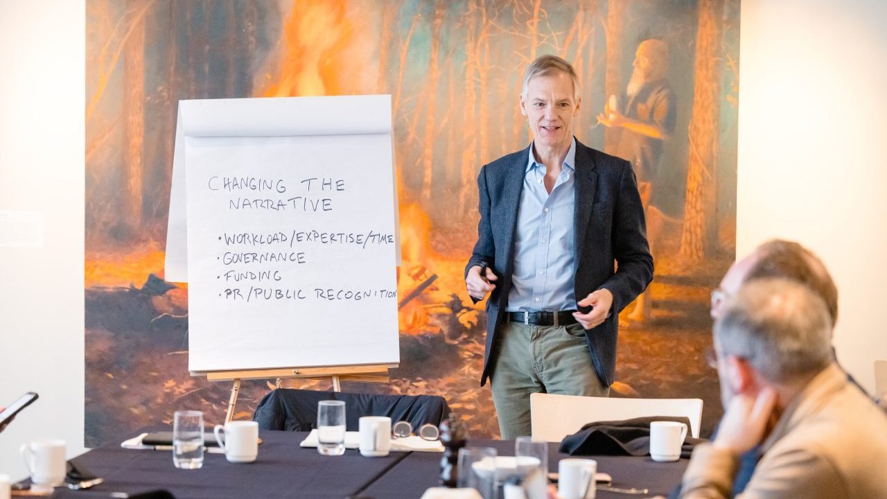 A person standing in front of a flipboard reading "Changing the Narrative: Workload/expertise/time, governance, funding, and PR/public recognition."