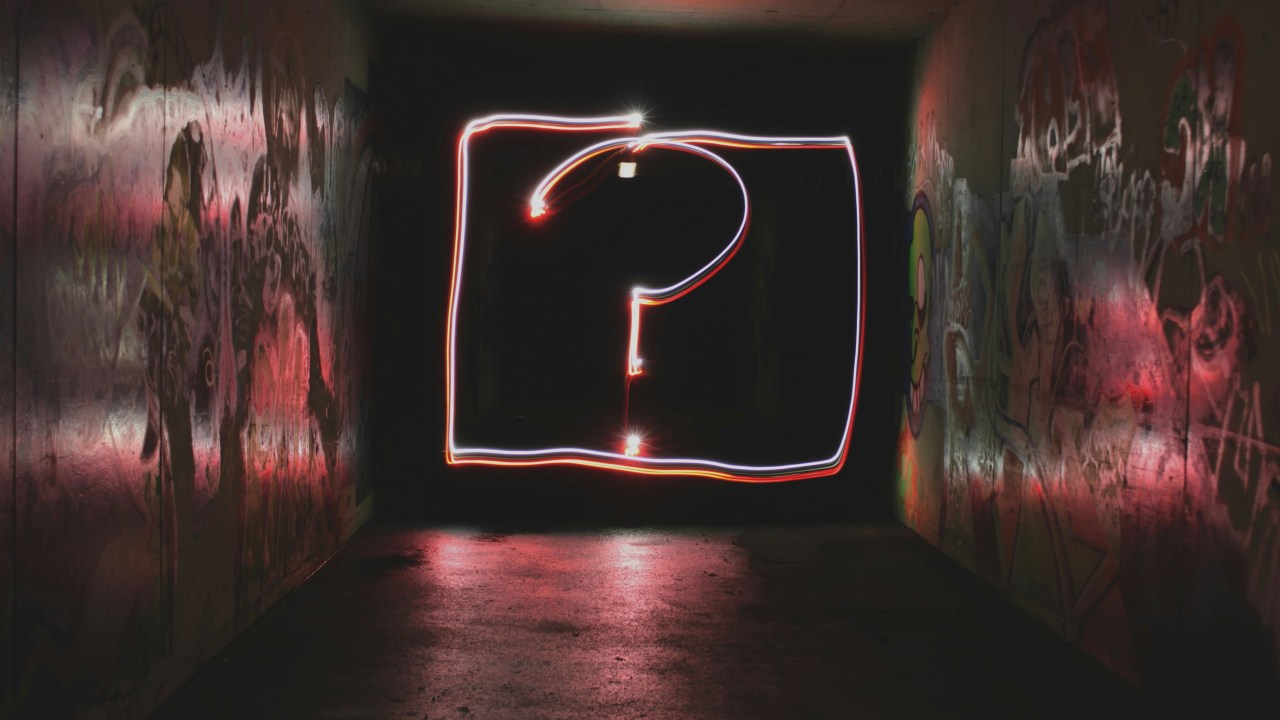 A dark tunnel with graffiti-covered walls and red lighting. A glowing question mark, created with light painting, floats in the center.