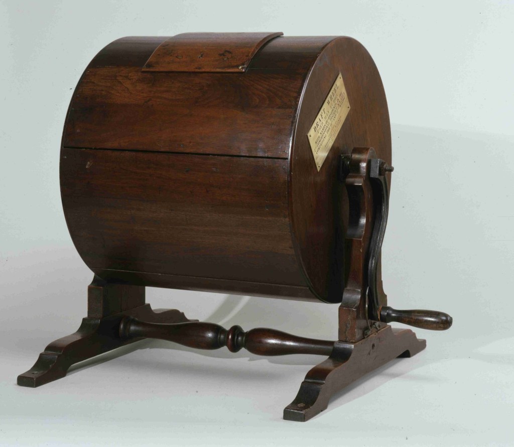 A wooden structure with a round barrel situated on top of a trestle with a wooden crank and an opening in the top of the barrel