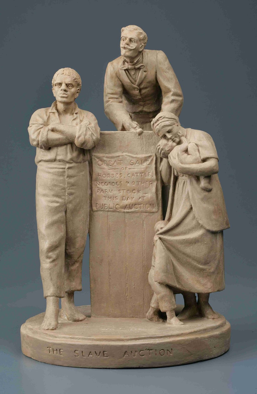 White marble sculpture with two men standing in front of and behind an auction block with a woman standing to the right hand side carrying a baby that she is kissing in a longing way. 