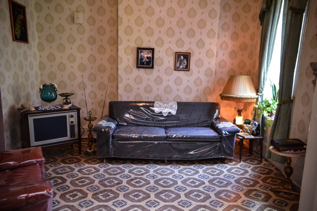 View of the interior of one of the Tenement Museums exhibits with a couch covered in clear plastic sitting on a patterned rug with a side table and lamp to the right and a 1960s television to the left. The walls have a patterned wallpaper.
