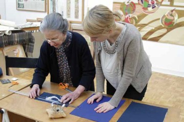 Two women stand at a ttable measuring fabric with a set of pins and other sewing tools around. 