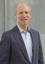 A balding white man stands smiling at the camera wearing a grey colored suit jacket and light blue button down shirt.