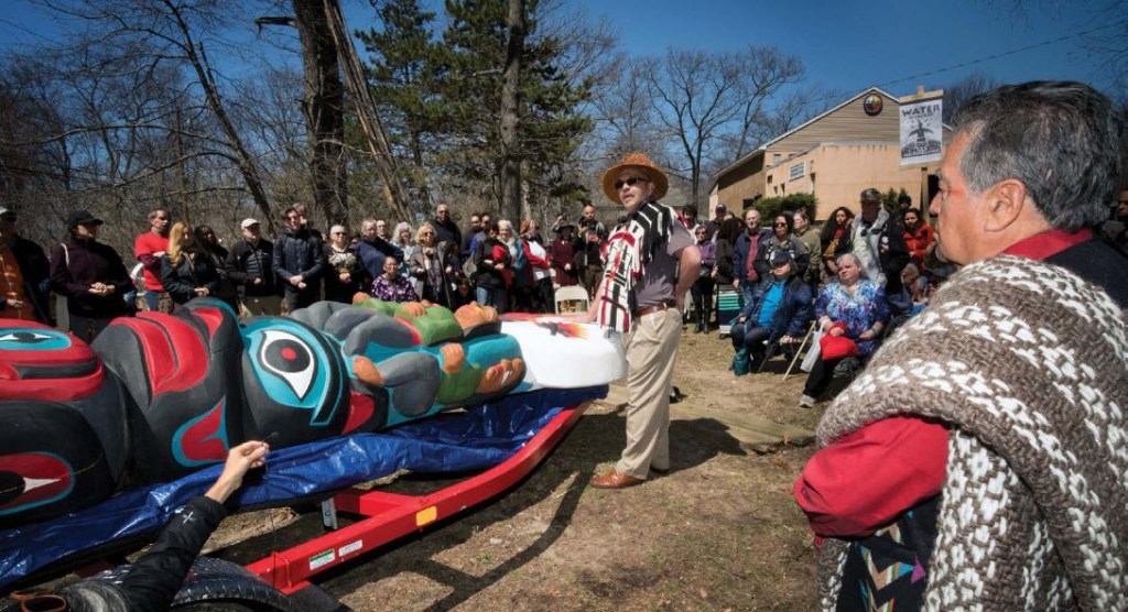 A group of people stand watching a two native men start to carry a colorful totum pole laying down off a trailer is wheeled past.