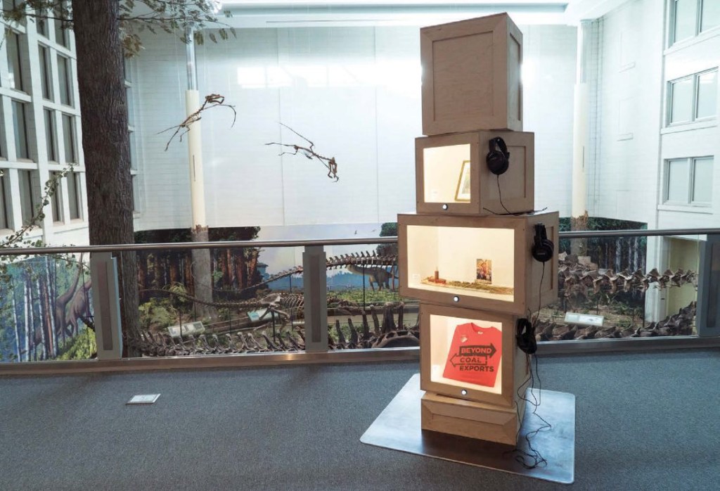 View of the interior space of an exhibition with what looks like a stack of crates with lighted interiors and headphones hooked to the side. 