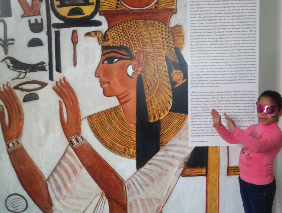 A young girl mimicks the Egyptian hygroglyph depicted in a large wall panel in a museum exhibition.
