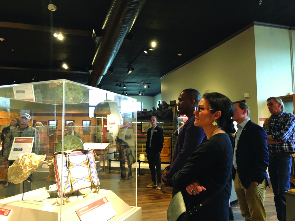 A group of people stand in a large gallery, one smaller group of two look at an object in an enclosed case in the foreground.
